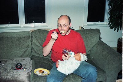 Man in red shirt brushing teeth while holding infant, seated on a vintage-style sofa. Yellow plate with food on armrest. Indo...