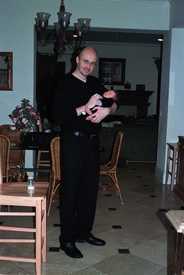 A man in formal attire holds a baby indoors, likely a 1980s-1990s dining/living space. Wooden furniture, floral arrangements,...