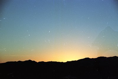 Silhouetted mountain range at dusk with streaks of light pollution and faint meteor trails against a gradient sky (blue to or...