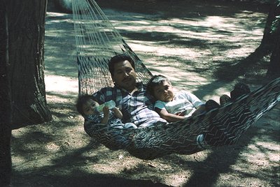 Vintage family moment in a woven hammock under dappled sunlight, mid-20th century. Adult and two children relax in a shaded f...