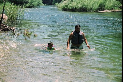 Adult guides young child in shallow river, surrounded by lush greenery. Reflective water suggests midday lighting. Likely 199...