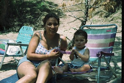 A woman and child relax on vintage beach chairs by a waterfront, likely mid-20th century. The woman holds a pink towel, while...