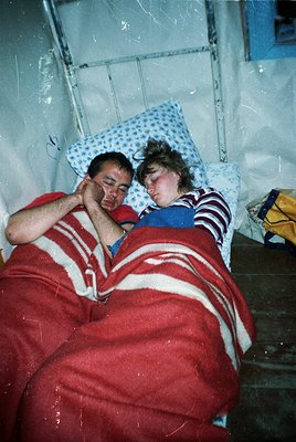 Two individuals wrapped in red-and-white striped blankets rest closely on a narrow bed in a dimly lit room. The setting sugge...
