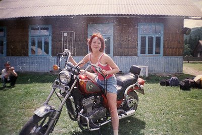 Vintage motorcycle with red/orange paintwork and chrome detailing, parked in front of a rustic wooden house with blue-framed ...