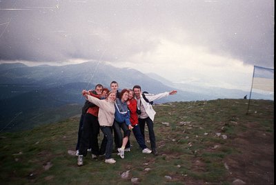 Vintage group photo on a grassy hilltop with mountain backdrop, 1990s-era clothing (tracksuits, sneakers). Five individuals p...