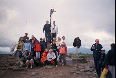 Group photo atop a mountain summit, likely in the 1990s. 15+ hikers pose on rocky terrain with a flagpole and cross marker. C...