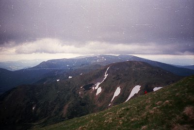 Vintage alpine landscape with snow patches on rocky terrain under low-hanging clouds. Single red backpacker visible mid-hike,...