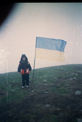 Vintage black-and-white photo of a child holding a Ukrainian flag on a windswept hillside, wearing a red backpack. Distressed...