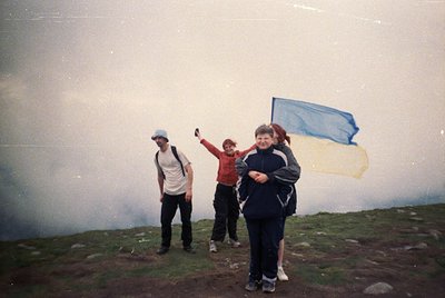 Three hikers pose atop misty mountain peak, 1990s-era gear visible. Blue flag waves in wind, suggesting summit achievement. F...