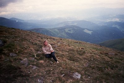 A lone individual sits atop a rugged, grassy alpine ridge, framed by rolling mist-covered valleys. The vintage filter suggest...