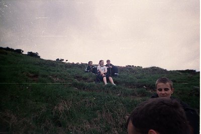 Vintage outdoor group shot on grassy hillside, likely 1980s–1990s. Four seated individuals in matching sportswear (dark jerse...