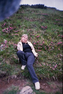 A woman in 1970s-style jeans, a black top, and a beige jacket reclines on a grassy hillside blanketed with pink wildflowers. ...