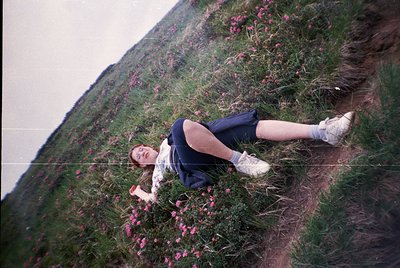Vintage candid of a person reclining on a grassy hillside amid blooming pink wildflowers, likely heather. Mid-20th-century ou...