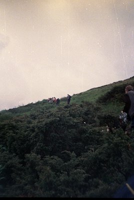 Vintage black-and-white photo of a group hiking on a steep, rocky hillside with sparse vegetation. Fog or mist obscures the u...