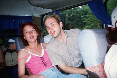 Vintage candid shot of two individuals in a moving vehicle, likely a bus or van, during the 1990s. The woman wears a sleevele...