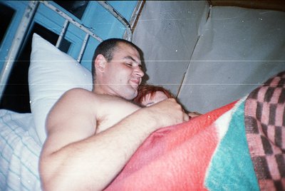 A man and child sleeping together on a bunk bed in a compact, utilitarian space. The bunk bed features a red blanket and a ch...