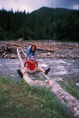 Two individuals pose on a fallen log near a flowing river, surrounded by forested hills. The man wears a red shirt and beige ...