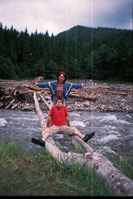 Two individuals pose on a fallen log over a shallow, rocky riverbed, surrounded by dense forest. The woman stands behind the ...