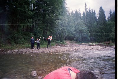 Vintage outdoor scene of three individuals playing frisbee near a shallow, rocky stream. Dense coniferous forest in backgroun...