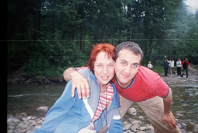 Couple posing by a shallow, rocky riverbank surrounded by dense forest. Woman in striped sweater and blue jacket, man in red ...