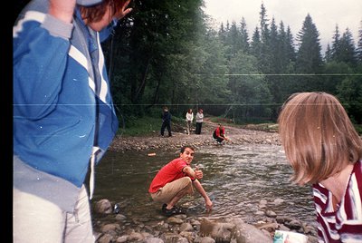 Vintage outdoor scene: group of people wading in shallow riverbed surrounded by dense coniferous forest. Central figure in re...