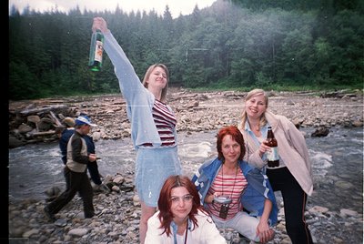 Vintage group photo by a rocky riverbank surrounded by dense forest. Four women pose with drinks, mid-1990s Eastern European ...