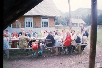 Group picnic in rustic alpine setting, 1990s. Wooden cabin with corrugated roof and metal-framed windows in background. Long ...