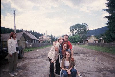 Vintage group photo in rural setting, likely Eastern Europe, 1970s-1980s. Four adults pose in front of wooden houses and a tr...