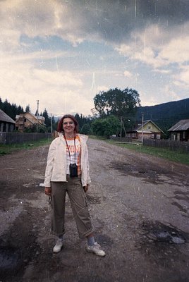 Vintage photograph of a woman in a rural village setting, likely Eastern Europe, 1970s–1980s. She wears a light jacket, patte...