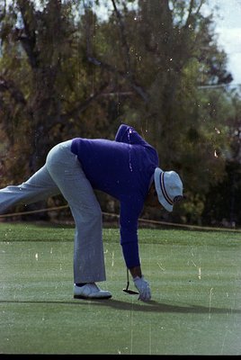 Mid-20th-century golfer in classic attire—light gray trousers, blue sweater, and white cap—executing a putt on a lush green c...