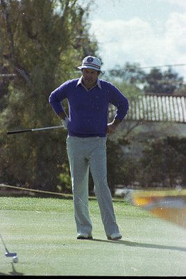 Mid-1970s golf scene: Man in vintage attire—white cap with logo, purple sweater, light trousers—mid-swing on a lush green fai...