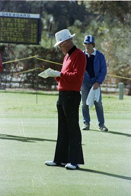 Mid-20th century golf course scene: Man in white fedora, red sweater, and dark trousers consults a scorecard on artificial tu...