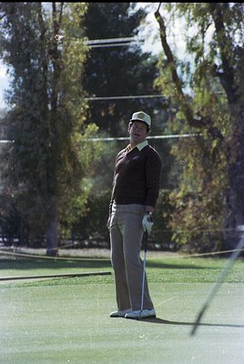 Mid-20th century golfer in vintage attire—light-colored cap, dark sweater, and slacks—poised on a lush green fairway. Golf cl...