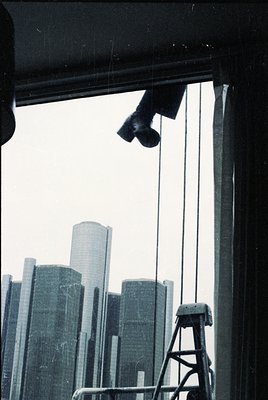 Urban skyline captured through a window with industrial lighting rigs. Mid-century modern high-rises dominate the background,...