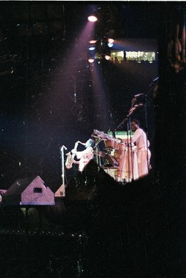 Vintage concert stage shot featuring a saxophonist in mid-performance, bathed in dramatic stage lighting. The musician wears ...