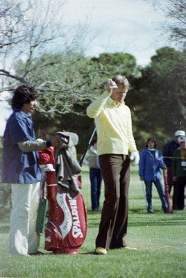 1970s golf scene featuring a golfer mid-swing in light pastel attire, red golf bag with "PGA" branding, and blurred spectator...