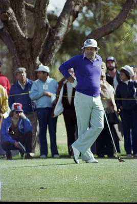 Mid-1970s golf scene: Player in vintage purple sweater and white cap mid-swing on a lush green fairway, surrounded by spectat...