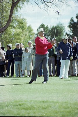 Mid-20th century golf scene: golfer in red sweater and white cap mid-swing on a lush green course, surrounded by spectators i...