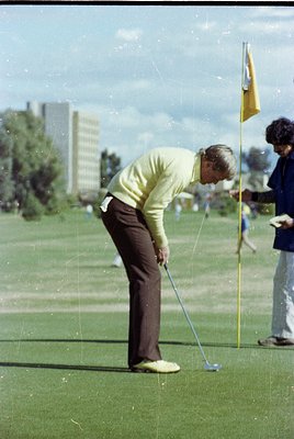 1970s golf scene: Male golfer in vintage sweater and trousers executes a putt on a lush green course. Urban high-rises and sp...