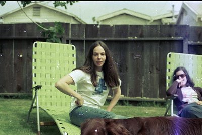 Vintage 1970s backyard scene: woman in long-sleeve graphic tee and bell-bottoms poses on a lime-green plastic lounge chair, w...