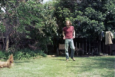Man in 1970s-style casual wear (red T-shirt, rolled jeans) tosses frisbee in a lush backyard. Dog mid-air captures motion. Wo...