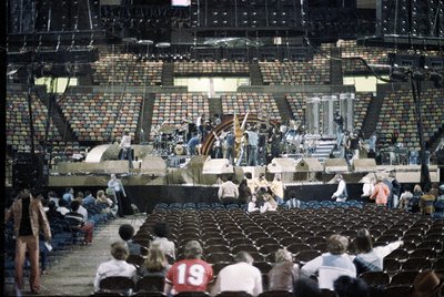 Vintage stadium interior with tiered brick seating and stage setup, likely for a concert or event. Crowd in front rows wearin...