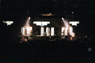 Vintage concert stage bathed in spotlight beams, featuring a drum kit and keyboardist silhouetted against dim lighting. Stage...