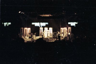 Vintage concert stage setup with spotlighted performers on raised platforms. Visible instruments include drums, keyboards, an...
