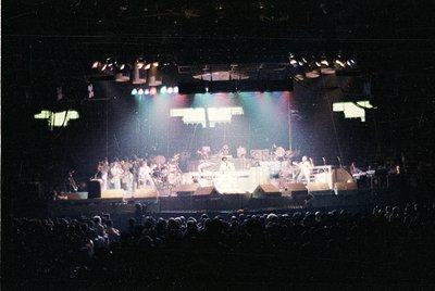 Vintage concert stage featuring a large band performing under colorful stage lights, likely 1970s–1980s. Drummer, keyboardist...