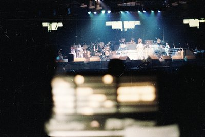 Vintage concert stage with band performing under blue stage lighting. Visible instruments include drums, keyboards, and guita...