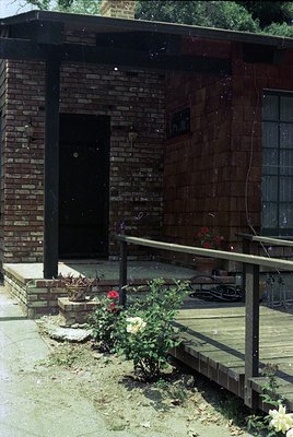 Mid-century brick entrance with concrete steps and wrought-iron railing, featuring a small landscaped garden with roses and p...