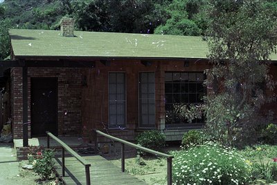 Mid-century modern brick home with green shingle roof, set in lush greenery. Brick foundation, large front windows, and a sma...