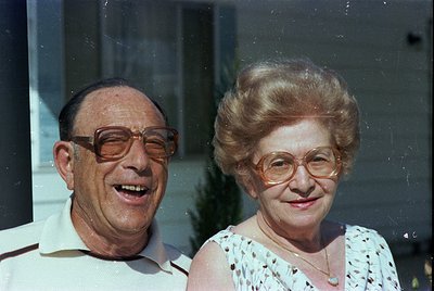 Retro portrait of an elderly couple posing indoors, likely mid-20th century (1960s–1980s). Man wears round, wire-framed glass...