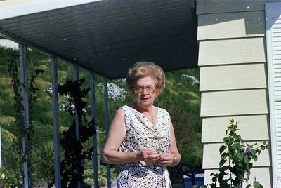 Mid-century woman in a floral dress poses under a covered patio, holding a small object. Light-colored wooden house with vert...